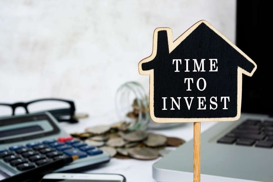 Un panneau en forme de maison avec l'inscription "Time to invest", posé sur un bureau près de pièces de monnaie et d'une calculatrice pour illustrer l'investissement immobilier.