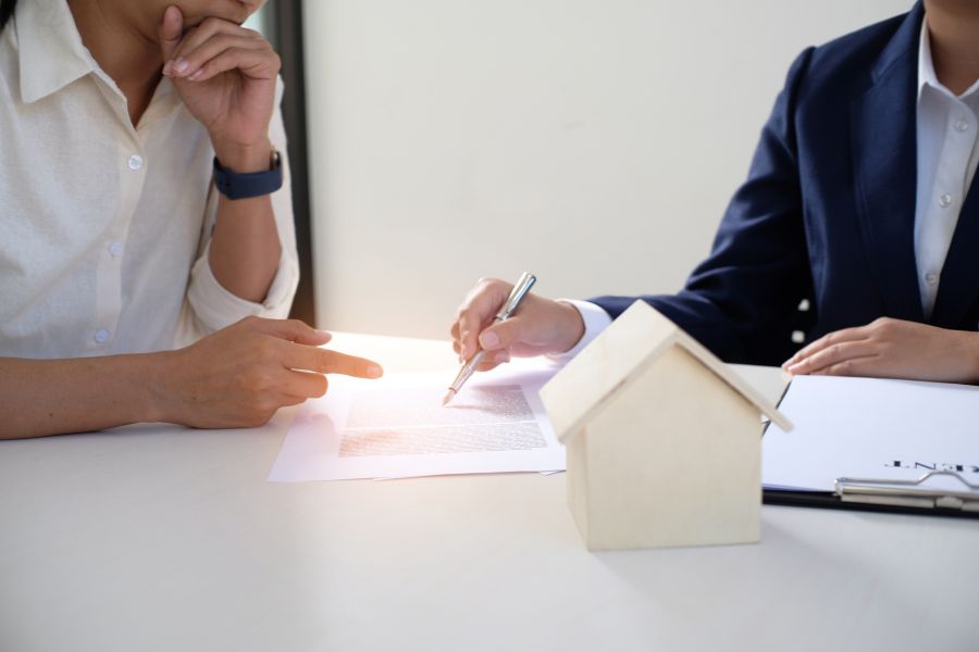 Un client et un agent d'assurance discutent des termes d'un contrat multirisque pour une maison, illustré par une maquette en bois.
